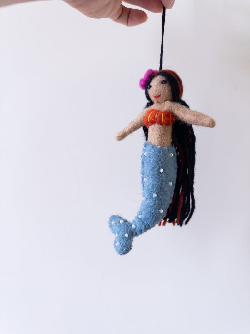 A handmade wool felt mermaid ornament is suspended by a hand against a plain white backdrop. This unique Christmas tree ornament features a tan upper body, a red top with yellow stripes, and a blue tail with white dots. The felt mermaid has long black hair with red strands and a pink flower on its head, adding a playful and colorful accent.