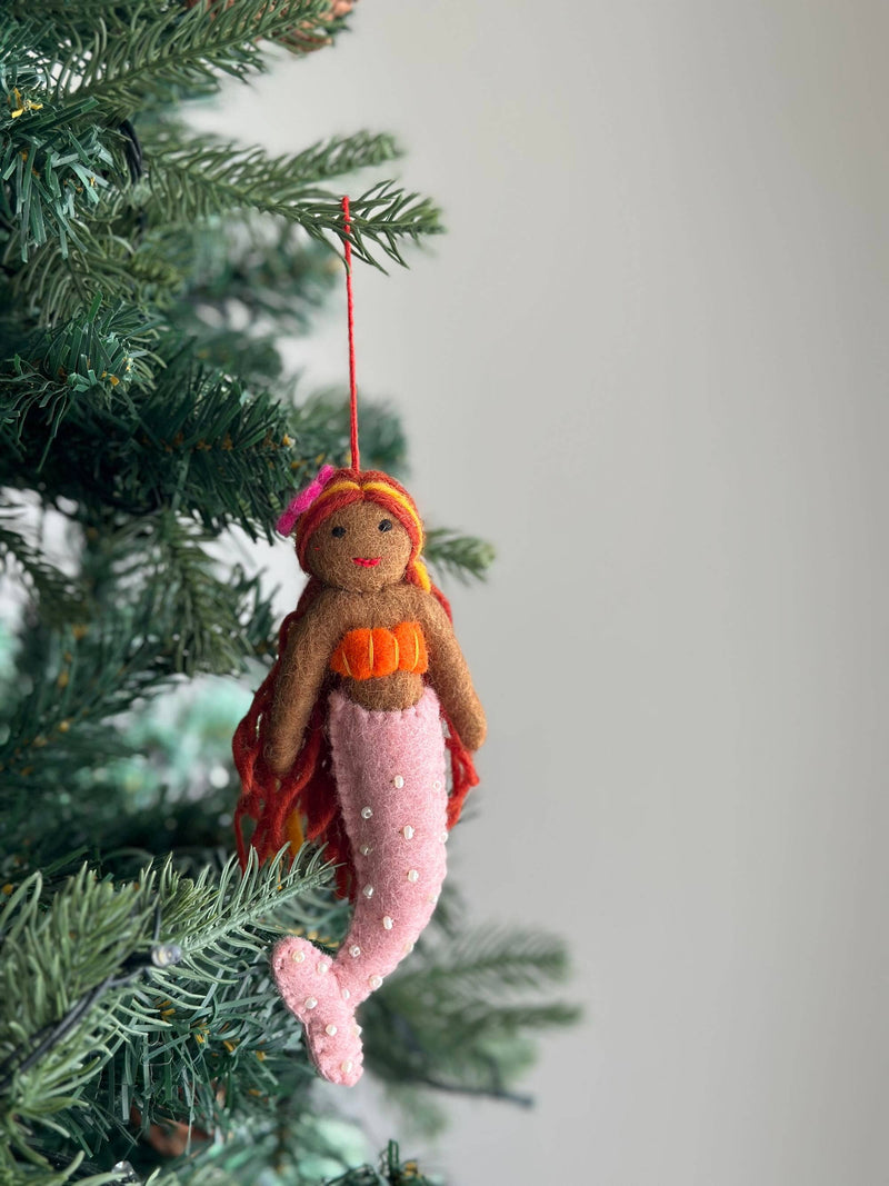 A whimsical wool felt mermaid ornament hangs from a Christmas tree branch, adding a festive touch. The unique ornament has a brown upper body with an orange bra, flowing orange and red yarn hair, and a pink flower on her head. Her pink tail is adorned with small white pearls, suspended by a red string.