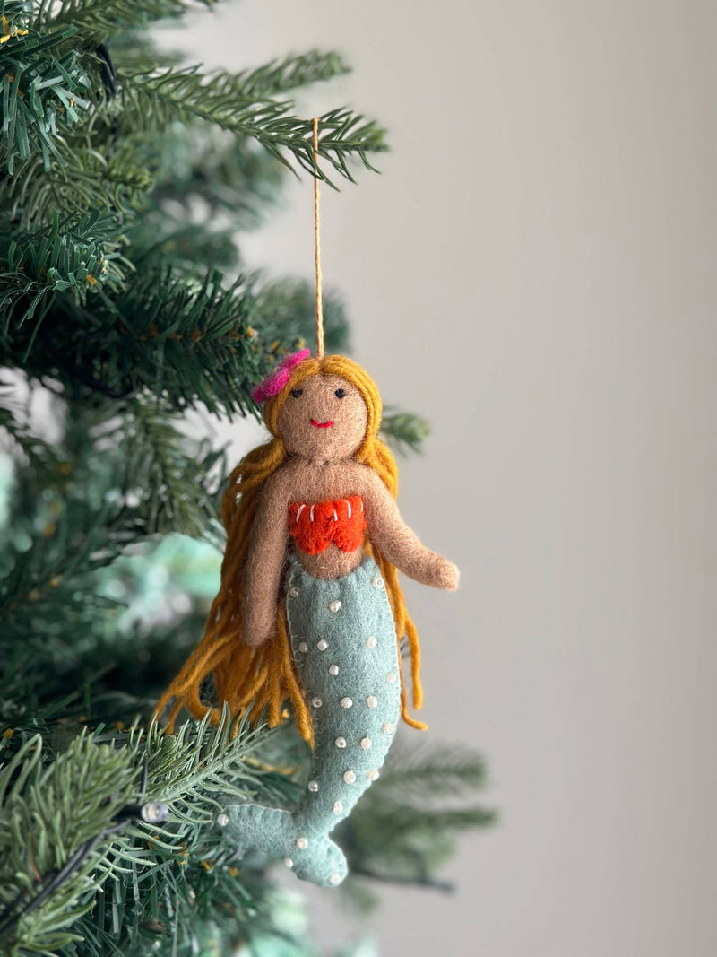 A playful wool felt mermaid Christmas ornament hangs on a vibrant green Christmas tree. This charming handmade ornament features light brown skin, long golden yellow hair adorned with a pink flower, a red shell bra, and a light blue tail embellished with white pearls, set against a soft, neutral background.