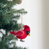 A red felt cardinal ornament hangs on a vibrant green Christmas tree, set against a bright, airy backdrop. This handcrafted felt cardinal features a red body with a yellow beak and intricate gray felt feet. Subtle white felt accents and a black felt accent around the eye enhance its textured body. A jute string allows for easy hanging.