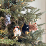 The four unique wool felt cat Christmas tree ornaments are displayed among green pine needles, adorning a lush Christmas tree in a close-up indoor shot with soft, warm light. Each of the 3D-designed, needle-felted cats has a jute string for hanging. From left to right, there's a gray tabby with dark gray stripes, a calico with black, white, and orange markings, a black cat with white whiskers, and a white cat.