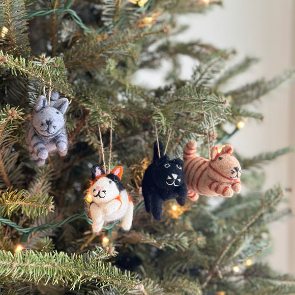 The four unique wool felt cat Christmas tree ornaments are displayed among green pine needles, adorning a lush Christmas tree in a close-up indoor shot with soft, warm light. Each of the 3D-designed, needle-felted cats has a jute string for hanging. From left to right, there's a gray tabby with dark gray stripes, a calico with black, white, and orange markings, a black cat with white whiskers, and a white cat.