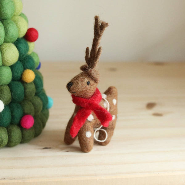 A festive wool felt reindeer Christmas ornament rests on a light wooden surface, positioned next to a decorative felt ball tree. This soft, handcrafted reindeer is brown with white spots, distinctive antlers, and a red scarf, featuring a white string loop on top.