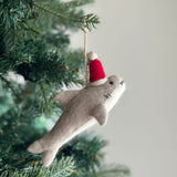 A festive Christmas shark felt ornament hangs on a lush Christmas tree, adding a playful touch to the holiday decor. This light gray shark ornament wears a red Santa hat with a white pompom. It features two black eyes and three black stitch marks for gills. A jute string is attached to the top for easy hanging.