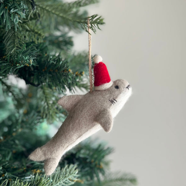 A festive Christmas shark felt ornament hangs on a lush Christmas tree, adding a playful touch to the holiday decor. This light gray shark ornament wears a red Santa hat with a white pompom. It features two black eyes and three black stitch marks for gills. A jute string is attached to the top for easy hanging.