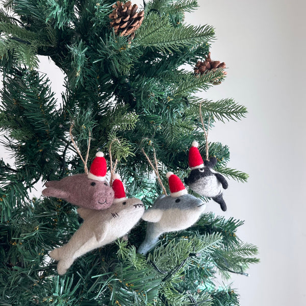 Felt Christmas shark ornaments hang festively on a green Christmas tree. Each handcrafted felt ornament features a rounded body, small fins, and stitched eyes, topped with a small red Santa hat and white pompom. The unique Christmas shark ornaments come in light gray with a white belly, darker brownish-gray, and black and white, each suspended by a jute string.