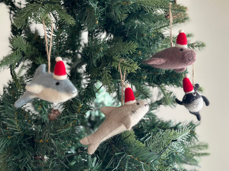 Multiple felt Christmas Shark ornaments hang gracefully on a lush green Christmas tree. Each handcrafted felt ornament is shaped like a shark, in varying shades of gray and brown, and adorned with a festive red and white Santa hat, complete with a jute string for hanging.