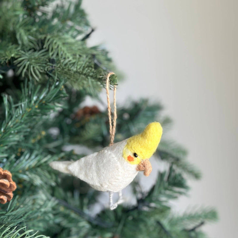 A handmade felt cockatiel Christmas ornament hangs delicately from a vibrant green Christmas tree. This charming felted cockatiel features a prominent yellow crest, a soft white body, and distinct orange cheek spots, capturing the bird's natural charm. It is suspended by a rustic jute string, adding a handcrafted touch.