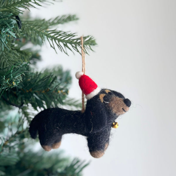 A charming handmade felt dachshunds ornament rests against a cheerful, festive backdrop, evoking a cozy and joyful atmosphere. This unique Christmas ornament showcases four adorable felt dachshunds in cream, khaki, brown, and black, encircling a playful felt bone. All elements are set on a white felt frame, complete with a rustic jute string for easy hanging, adding puppy charm.