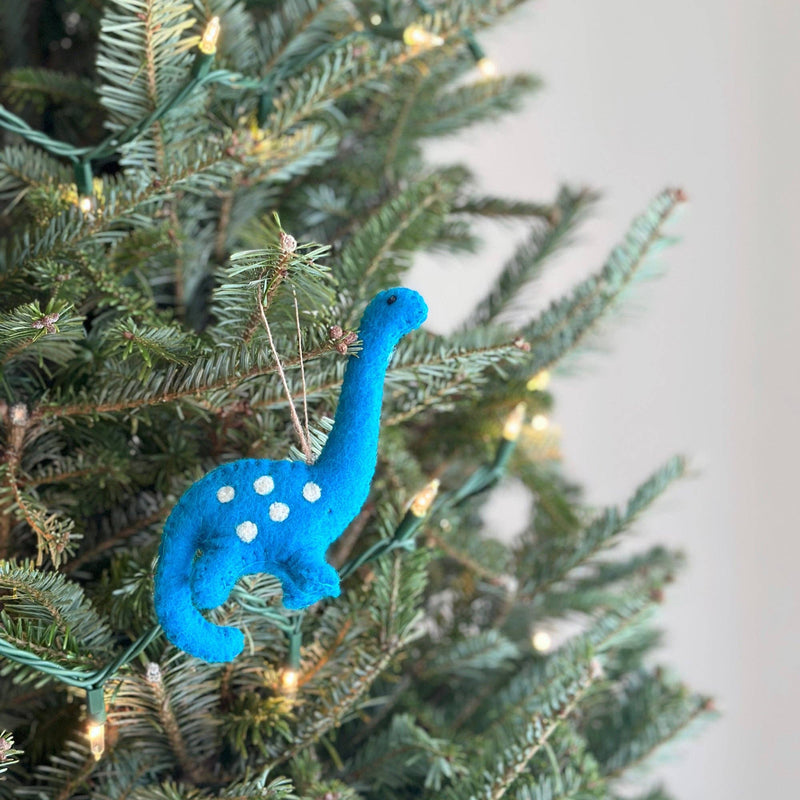 A playful blue wool felt brontosaurus ornament hangs on a lit Christmas tree. This handmade felt brontosaurus ornament features a long neck, a rounded body, small legs, and white polka dots. A jute string is attached for easy hanging, adding a festive touch to holiday decor.