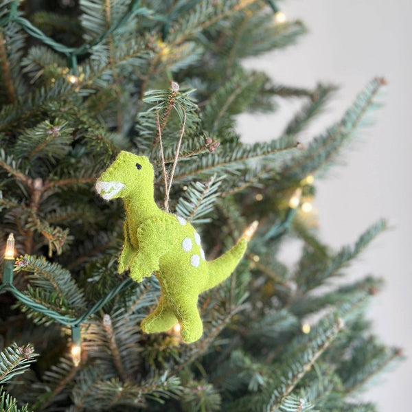 A light green wool felt T-rex Christmas ornament hangs from a Christmas tree adorned with warm, small lights, creating a festive atmosphere. This unique T-rex felt ornament features its distinctive shape with small arms and white spots, highlighting its textured surface and a jute string for hanging.
