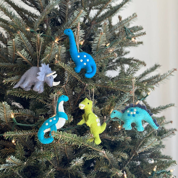 Hand-stitched wool-felt dinosaur ornaments, including a green T-rex, are displayed on the branches of a Christmas tree with warm white lights, creating a joyful mood. The collection features various unique felt dinosaur ornaments: a gray triceratops with white horns, blue and light blue brontosauruses with white dots, and a blue stegosaurus with dark blue accents, each with a jute string for hanging.