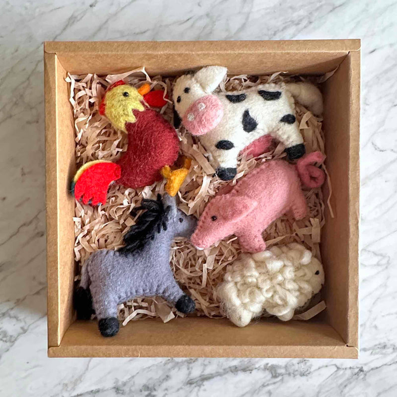 A handmade felt donkey ornament is presented inside a square cardboard box filled with brown paper shred, resting on a mottled white surface. The wool felt donkey ornament features a gray body with a black mane and hooves, showcasing a distinct wooly texture. This charming Christmas tree ornament evokes a rustic and playful mood, ready for gifting or display.