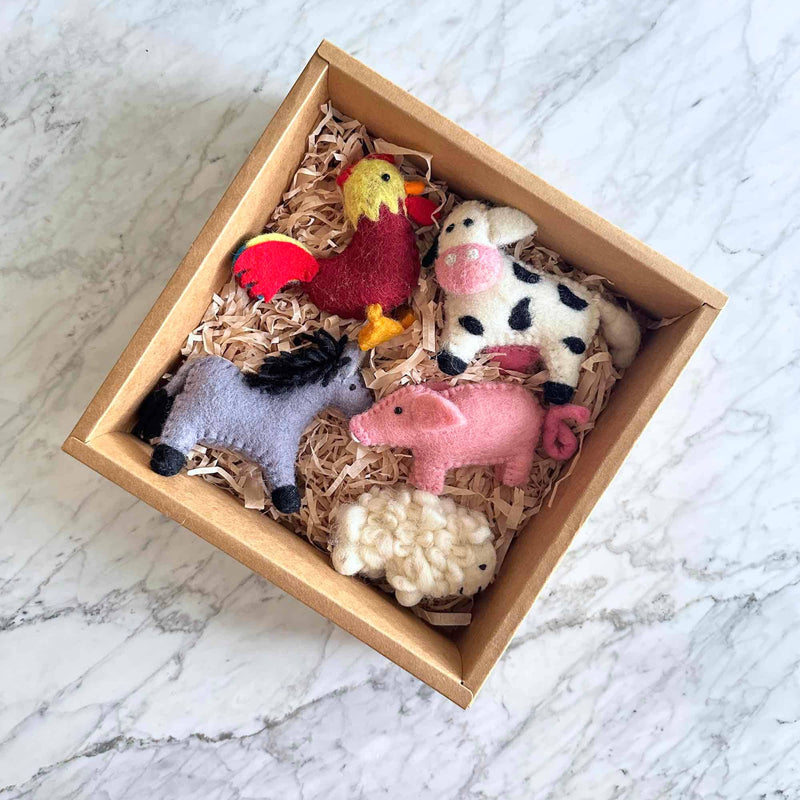 A light purple wool felt donkey ornament with a black mane rests inside a square cardboard box filled with shredded paper, positioned on a white marble surface. This unique handmade felt ornament features distinctive textures and patterns, reflecting its charming handcrafted design, perfect for decorating or collecting.