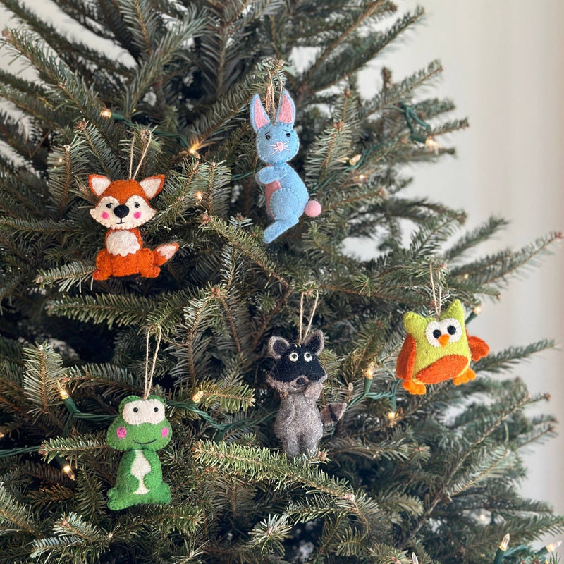 A handmade felt frog ornament hangs on the branches of a Christmas tree adorned with small, warm lights. The green frog ornament features pink cheeks and is suspended by a jute string, adding a rustic and unique handcrafted touch to the festive display.