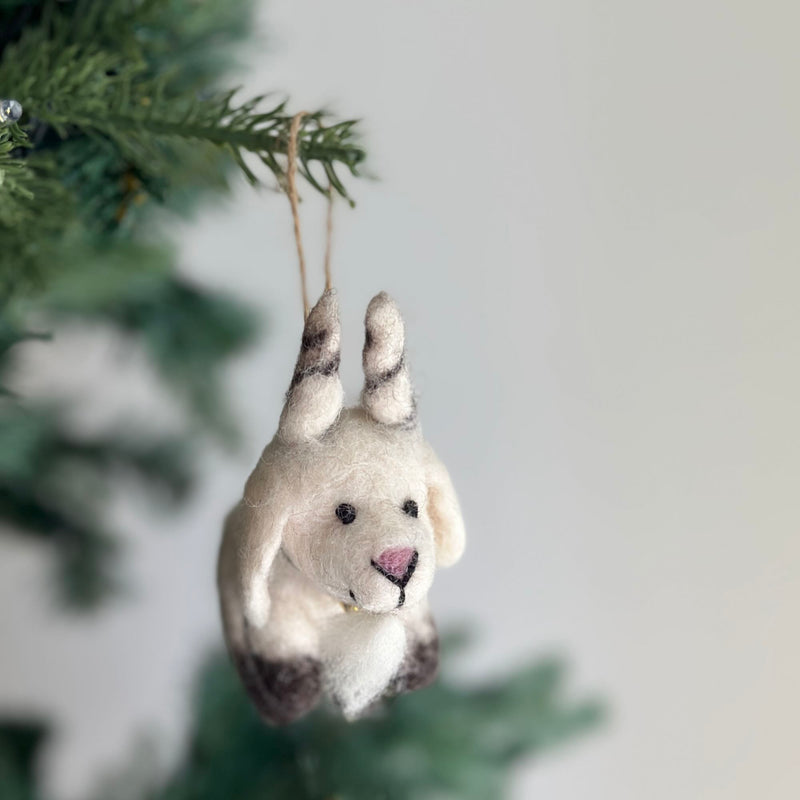 A needle-felted organic wool goat Christmas ornament hangs from a Christmas tree branch, showcased against a soft, blurred white background. The off-white goat ornament features dark brown patches on its feet and ears, a pink triangular nose, and small black eyes, suspended by a rustic jute string.