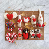 A felt Graduation Black Bear ornament, holding a heart, is displayed on a woven mat against a white marble backdrop. This handcrafted Christmas ornament features a black bear in a graduation cap and gown clutching a small red heart. Made from felt, it includes a jute string for hanging, bringing a whimsical touch to any display.