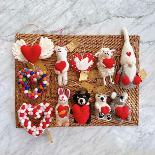 A felt Graduation Black Bear ornament, holding a heart, is displayed on a woven mat against a white marble backdrop. This handcrafted Christmas ornament features a black bear in a graduation cap and gown clutching a small red heart. Made from felt, it includes a jute string for hanging, bringing a whimsical touch to any display.