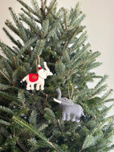 A light gray felt elephant ornament hangs from a lush Christmas tree branch by a jute string. This unique Christmas ornament features a detailed texture, a long trunk, large ears, and a darker gray tail, adding a cozy, festive touch to holiday decor.