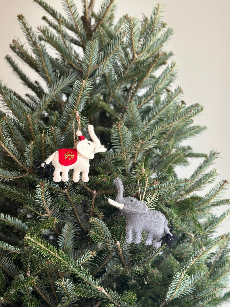 A light gray felt elephant ornament hangs from a lush Christmas tree branch by a jute string. This unique Christmas ornament features a detailed texture, a long trunk, large ears, and a darker gray tail, adding a cozy, festive touch to holiday decor.