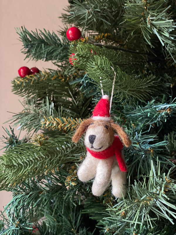 A cheerful wool felt Jack Russell dog ornament with a Christmas hat hangs on a festive Christmas tree adorned with red ornaments. This handmade felt ornament features a white body and brown ears, sporting a red Christmas hat with a white pompom and a matching red scarf. Its face has a black nose and stitched smile, with a white cotton string for hanging.