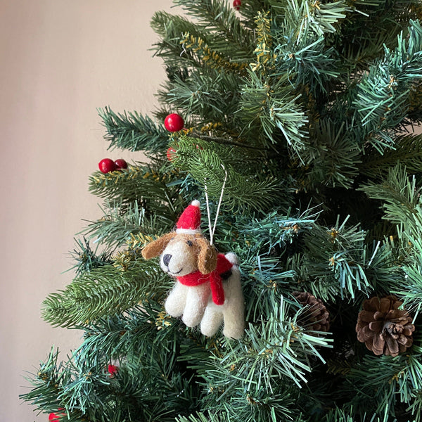 A charming wool felt Jack Russell dog Christmas ornament, adorned with a red hat and scarf, hangs on a festive green Christmas tree. This handmade felt ornament, crafted from white and brown organic wool, features a black nose, a cheerful smile, and floppy brown ears. The vibrant tree is decorated with red berries and brown pine cones.