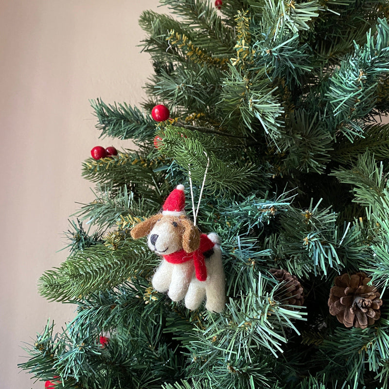 A charming wool felt Jack Russell dog Christmas ornament, adorned with a red hat and scarf, hangs on a festive green Christmas tree. This handmade felt ornament, crafted from white and brown organic wool, features a black nose, a cheerful smile, and floppy brown ears. The vibrant tree is decorated with red berries and brown pine cones.