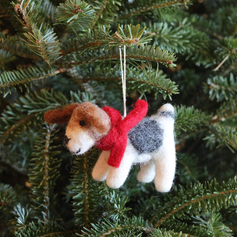A handcrafted Jack Russell dog felt ornament hangs by a white string from a vibrant green Christmas tree branch. This wool felt ornament features a predominantly white body with a dark grey spot on its back and a textured brown head. A bright red felt scarf is wrapped snugly around its neck, adding a festive touch.