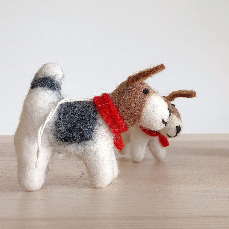 Two cheerful Jack Russell dog felt ornaments stand on a light wooden surface against a bright white background. The foreground needle felted ornament is a 3D Jack Russell dog Christmas ornament, crafted from white, black, and brown organic wool felt, wearing a vibrant red scarf. A white cotton string is attached for hanging, with a second similar Jack Russell dog felt ornament blurred behind it.