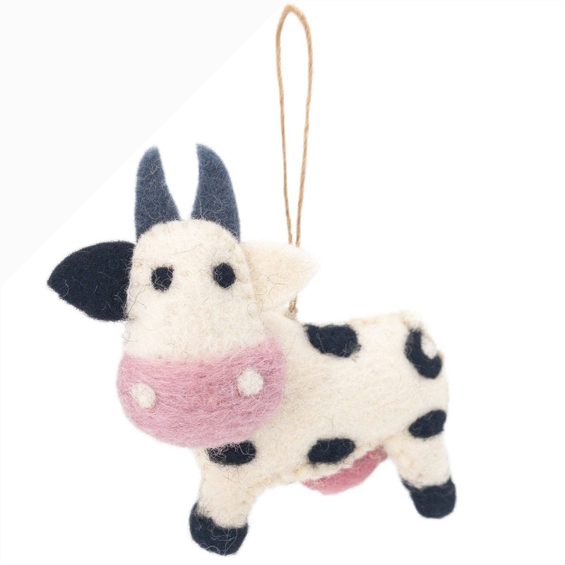 A handmade wool felt milk cow ornament is displayed against a plain white backdrop. This whimsical felt milk cow features a cream-colored body with black spots, blue horns, and a pink face. A jute string is attached for easy hanging.
