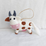 A handcrafted wool felt Milk Cow ornament is displayed against a bright white background. The charming felt cow has a white body with distinctive brown spots, brown hooves, and ears. Its face features small black eyes, gray horns, and a pink muzzle. A jute string is attached for easy hanging.