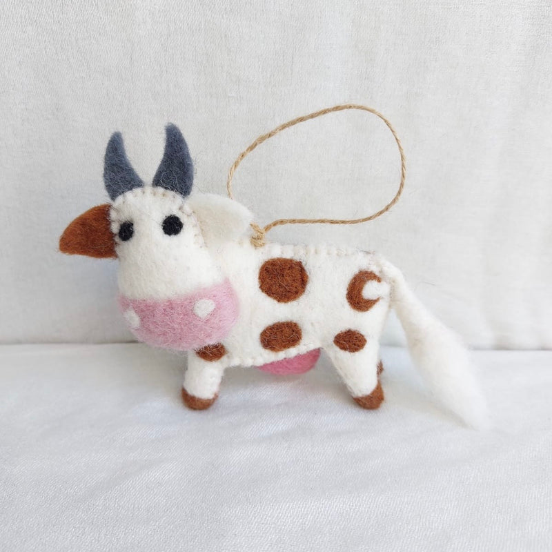 A handcrafted wool felt Milk Cow ornament is displayed against a bright white background. The charming felt cow has a white body with distinctive brown spots, brown hooves, and ears. Its face features small black eyes, gray horns, and a pink muzzle. A jute string is attached for easy hanging.