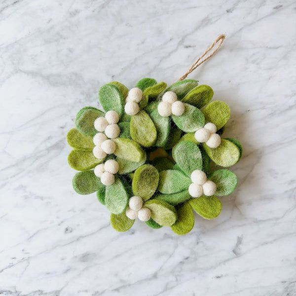 A mini felt mistletoe wreath ornament rests on a marble surface. This 4-inch unique Christmas ornament features a faded white tie-dye design on felt, circularly shaped on a rustic jute hoop, with a jute string for hanging.