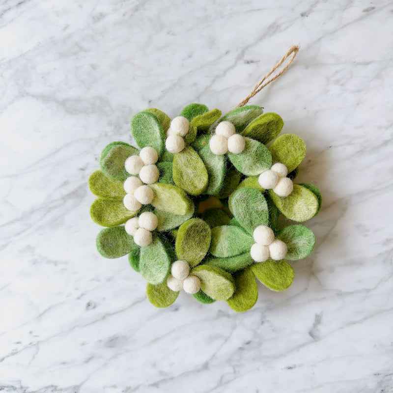 A mini felt mistletoe wreath ornament rests on a marble surface. This 4-inch unique Christmas ornament features a faded white tie-dye design on felt, circularly shaped on a rustic jute hoop, with a jute string for hanging.