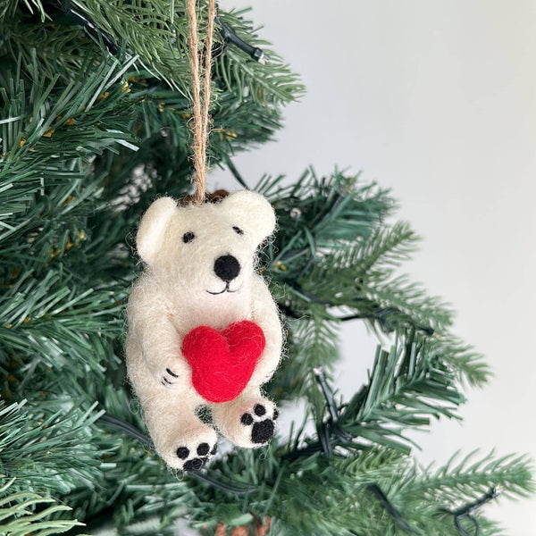 A charming felt mini polar bear ornament hangs on a Christmas tree, exuding a festive and joyful atmosphere. This white handmade felt ornament features black eyes, nose, and paw pads, and sweetly holds a vibrant red heart.