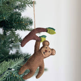 A brown felt monkey ornament hangs playfully from a lush Christmas tree branch, creating a joyful and festive atmosphere. Crafted from brown felt, the charming monkey appears to dangle from a matching felt branch adorned with light green felt leaves. It features a smiling face and small round ears, suspended by a jute string.