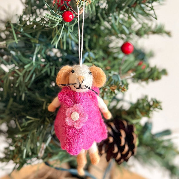 A needle felted mouse Christmas ornament hangs closely on a Christmas tree branch. This handcrafted felt ornament features a light cream mouse with tan ears, feet, and black whiskers. It wears a bright pink dress adorned with a round white flower, embodying its 'Girl' theme. Red berries and a pine cone decorate the festive tree.