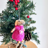 A charming felt mouse ornament, depicting a girl, hangs on a festive Christmas tree. The handmade mouse features a light beige head, long ears, and a black stitched nose. It wears a bright pink dress adorned with a white flower. A white cotton string allows it to hang among green needles, red berries, and a pine cone.