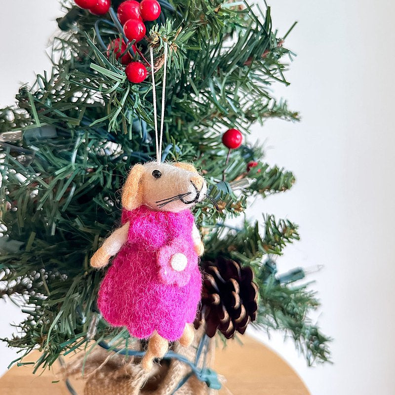 A charming felt mouse ornament, depicting a girl, hangs on a festive Christmas tree. The handmade mouse features a light beige head, long ears, and a black stitched nose. It wears a bright pink dress adorned with a white flower. A white cotton string allows it to hang among green needles, red berries, and a pine cone.
