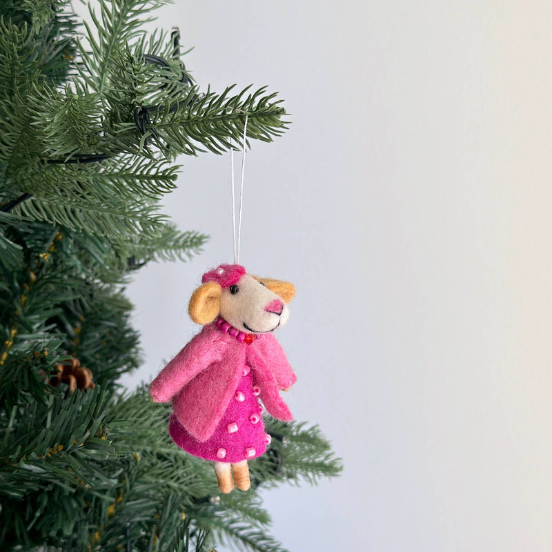 A charming handmade felt mouse ornament hangs on a vibrant green Christmas tree branch, with a pine cone visible. This felt mouse has a light beige face with round ears, a pink nose, and a red smile. It wears a pink beaded dress, matching jacket, hat, and a beaded necklace, suspended by a delicate cotton string.