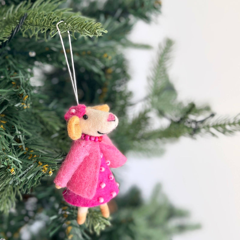 A charming wool felt mouse Christmas ornament hangs from a green Christmas tree branch, suspended by a silver string. The handmade mouse features a cream-colored head with rounded tan ears, a pink nose, and black eyes. It is adorned with a pink bow on its head and a matching beaded necklace, dressed in a pink dotted dress and a pink jacket.