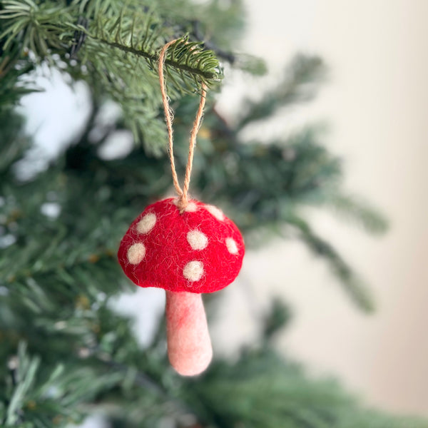 A cheerful felt mushroom ornament hangs from a green Christmas tree branch, suspended by a jute string. This handcrafted felt ornament features a bright red cap with white spots and a light pink stem, its soft, tactile texture adding a whimsical touch to holiday decor.