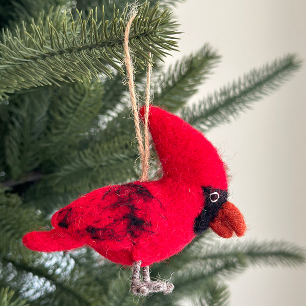 A needle felted cardinal ornament hangs gracefully on a Christmas tree. This bright red felt cardinal features a pronounced crest, small black markings on its back, a neatly shaped tail, and brown beak and legs. A jute string adds to its rustic appeal, perfect for festive charm.