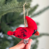 A needle felted cardinal ornament hangs from a jute string against a softly blurred Christmas tree background. This charming felt cardinal features a bright red body and head, accented with black markings, subtle black spots, black eyes, and a textured orange beak. It also has small gray felt feet.