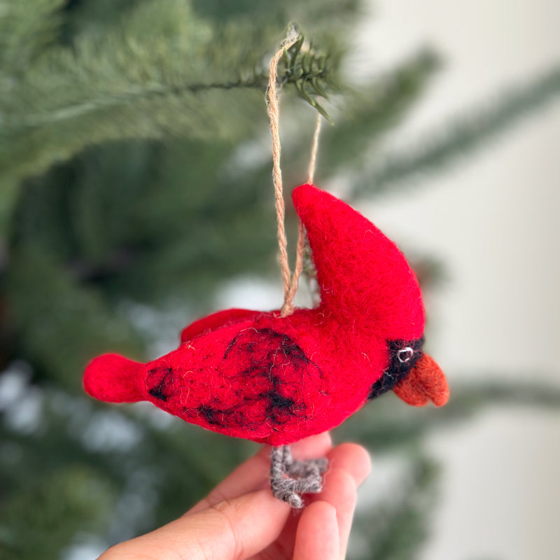 A needle felted cardinal ornament hangs from a jute string against a softly blurred Christmas tree background. This charming felt cardinal features a bright red body and head, accented with black markings, subtle black spots, black eyes, and a textured orange beak. It also has small gray felt feet.