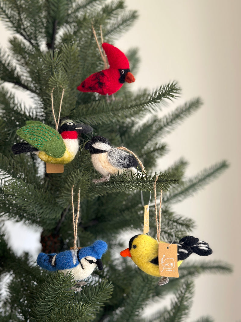 A needle felted cardinal ornament, crafted from red wool felt, hangs prominently on a Christmas tree. The cardinal features a black face and darker red speckles, suspended by a natural jute string that blends with the green needles. This handcrafted felt Christmas ornament adds a cozy, handmade charm.