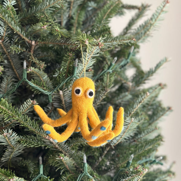 A handcrafted yellow octopus felt ornament hangs on a Christmas tree adorned with delicate lights. This vibrant yellow octopus features eight legs detailed with small blue dots and big white eyes with black pupils. A yellow cotton string is attached to its head, allowing it to hang gracefully, adding a cheerful holiday touch.