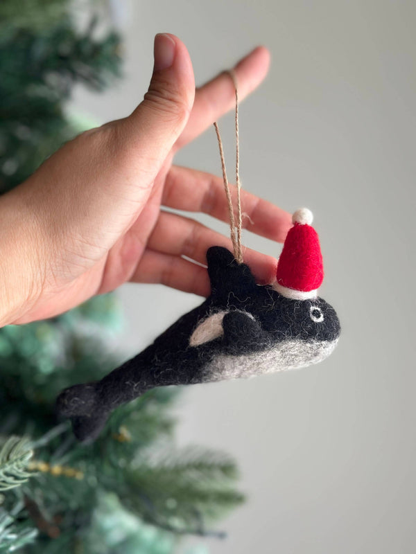 A playful felt orca ornament with a Christmas hat is held by a female hand against a blurred Christmas tree background. This charming needle felted orca ornament is primarily black with a white patch and a lighter gray underside. A whimsical red and white Christmas hat adorns its head, and a jute string is attached for hanging.