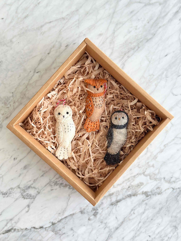 Felt owl ornaments from the 'Owl Family' collection are nestled inside a square cardboard box filled with shredded paper, set on a calm white marble surface. These handmade felt owl ornaments come in white with black speckles, brown with intricate stitching, and grey with a contrasting dark grey face and feet. Each unique Christmas ornament is attached with a red and white cotton string, suggesting it's ready for display.