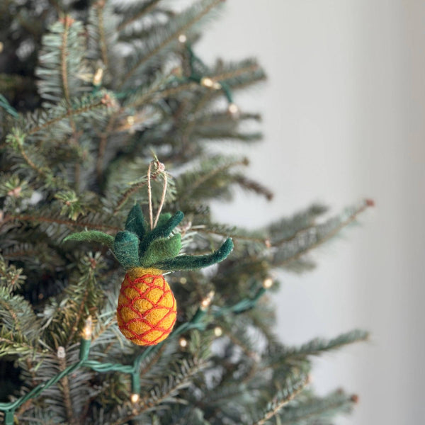 A handmade felt pineapple ornament hangs gracefully from a pine Christmas tree, illuminated by soft lights. This felt pineapple ornament features a textured yellow body with red crisscross detailing. It is topped with a vibrant array of green felt leaves, and includes a jute string for easy hanging.
